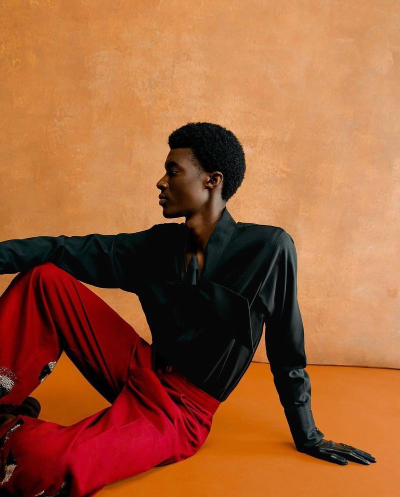 A person sits indoors on an orange surface, modeling in a black long-sleeved shirt, black gloves, and flowing red pants against a textured orange wall.