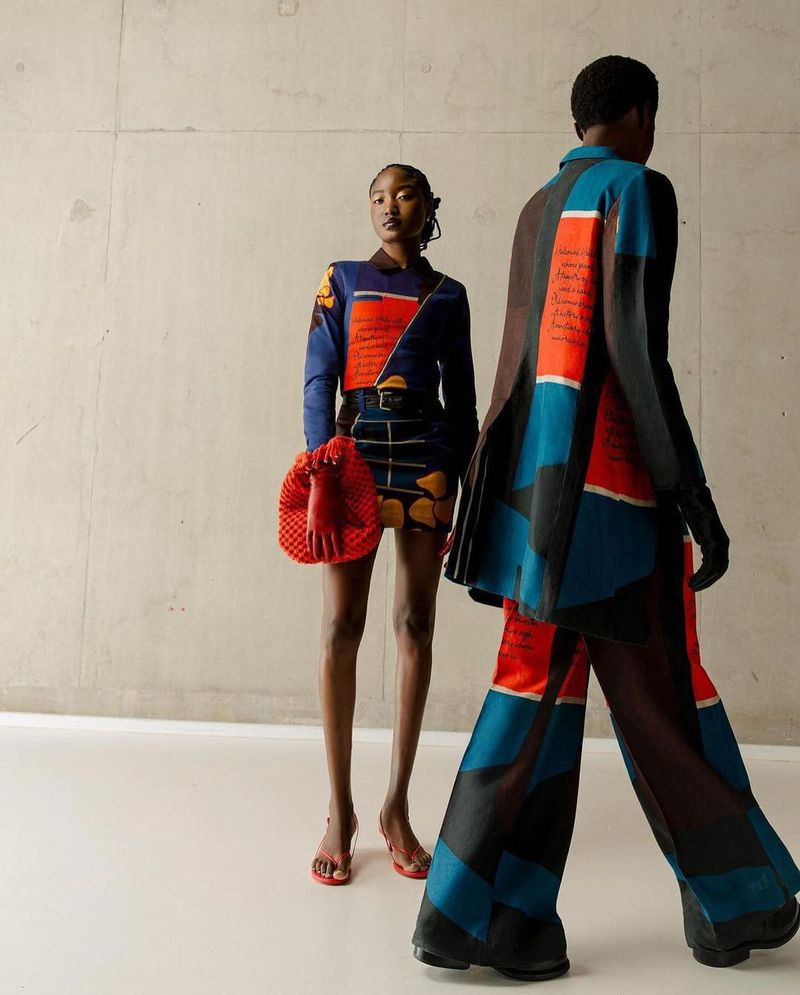Two Black fashion models showcase patterned outfits, one with a red woven bag, against a neutral backdrop in an indoor studio shot.