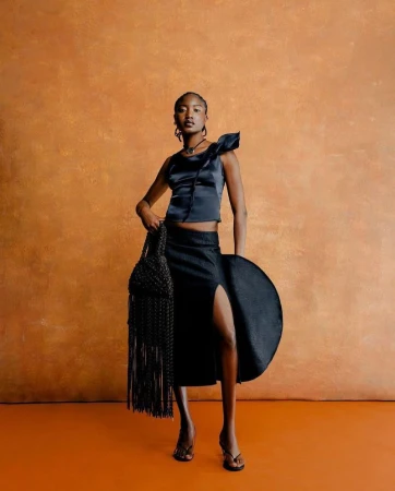 Full-length portrait of a Black woman in a dark blue top and skirt, holding a woven bag and disc, against an orange background.