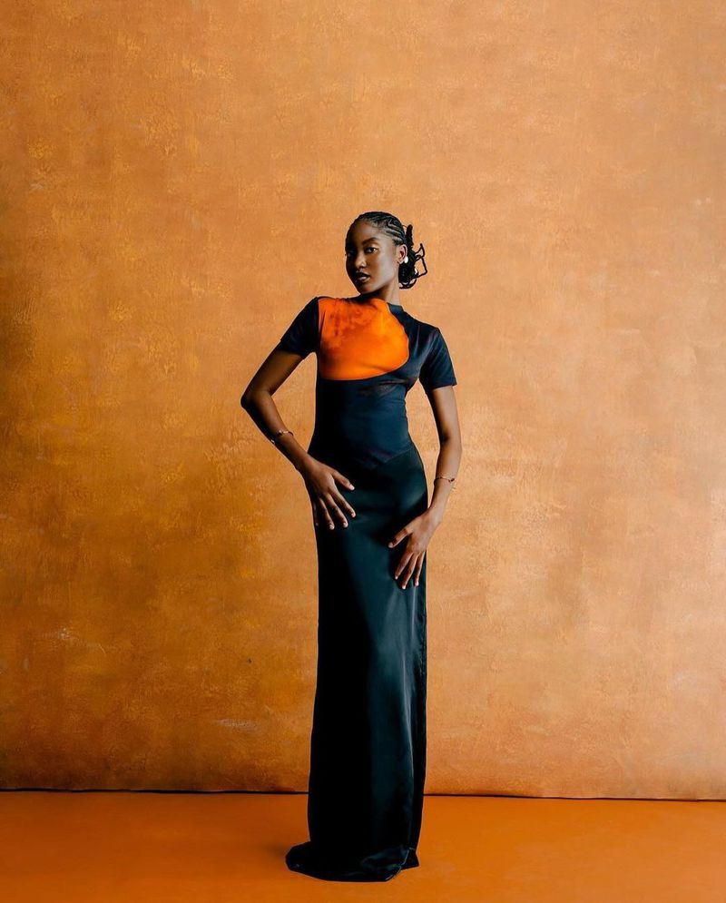 An elegant black woman stands gracefully in a floor-length black and orange dress against an orange studio backdrop in a medium shot.