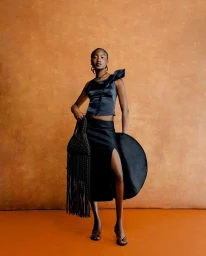 Model wearing a black satin top and slit skirt holding a fringed bag against an orange backdrop