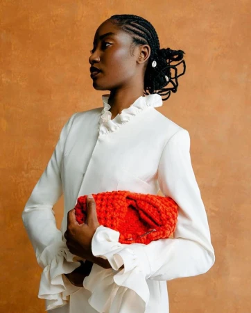 Black woman in white ruffled top holding a bright orange woven clutch against textured warm orange backdrop