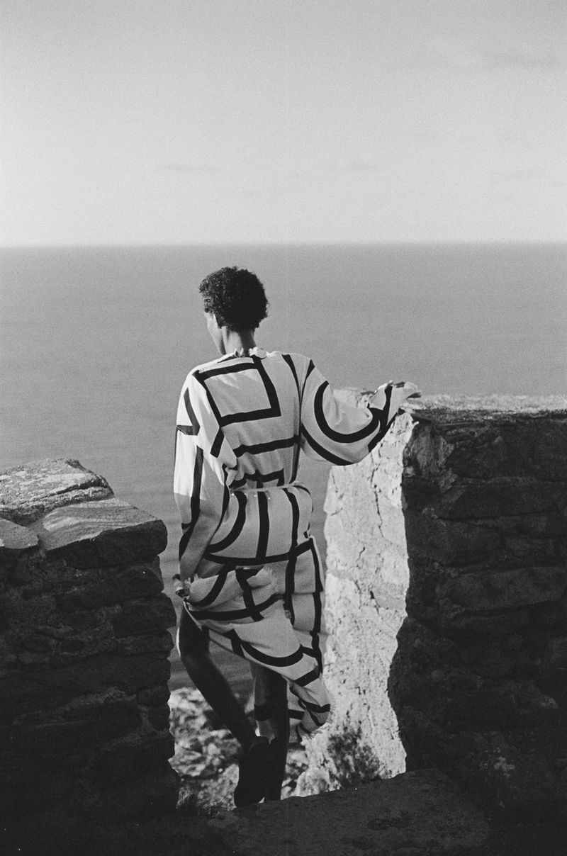 Black and white photograph of a person standing on a stone ledge overlooking the ocean, wearing a patterned robe. Minimalist composition.
