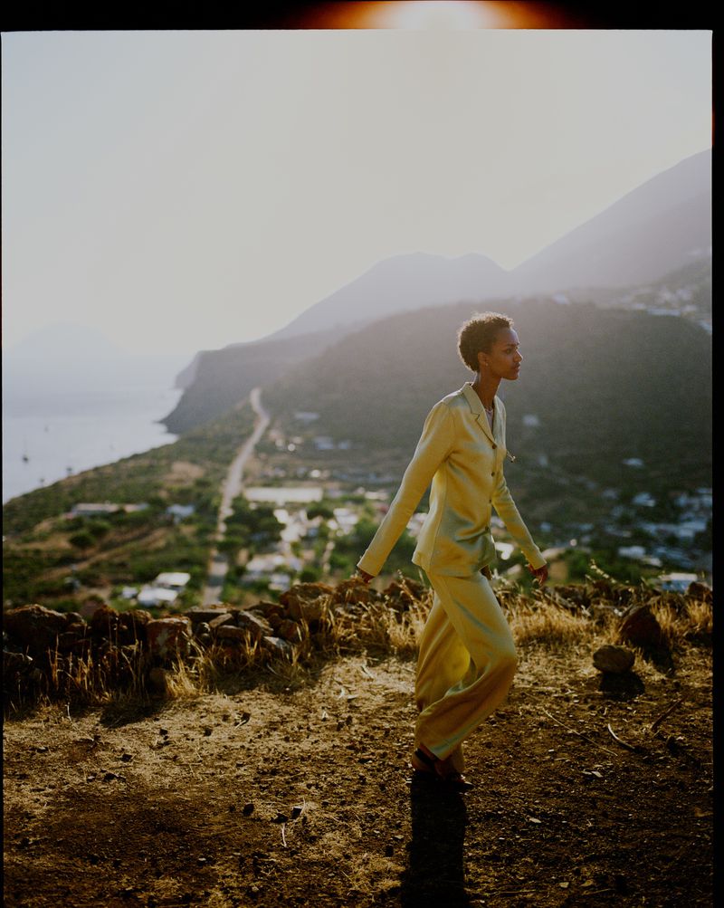 Woman in a yellow suit walks along a hilltop at sunset with mountains and the ocean in the background, creating a warm, outdoor fashion moment.