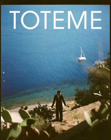 Adult in suit stands on coastal path overlooking ocean with a sailboat on the water and Mediterranean vegetation in the foreground.