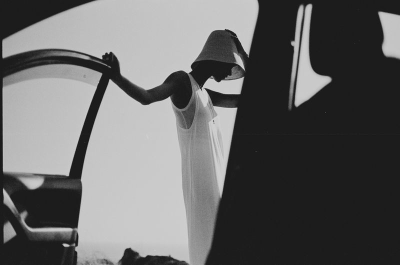 Monochrome portrait of a woman in a white dress and sunhat, leaning against a car door with a bright sky backdrop, creating stark shadows.