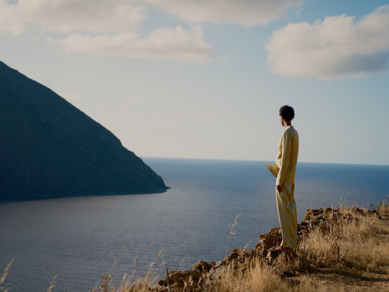 Adult stands on grassy hilltop overlooking dark blue sea water under a pale blue sky near a dark green mountain.