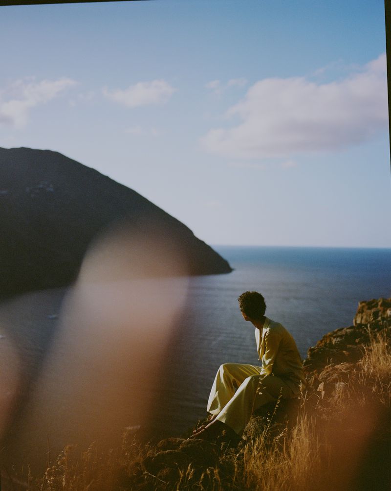 Adult male in pale yellow outfit sitting on a cliff overlooking a blue ocean with an island in the distance, against a light blue sky.