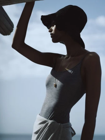 A woman wearing a gray swimsuit and black sun hat stands in front of a blue sky. She has a towel wrapped around her waist.