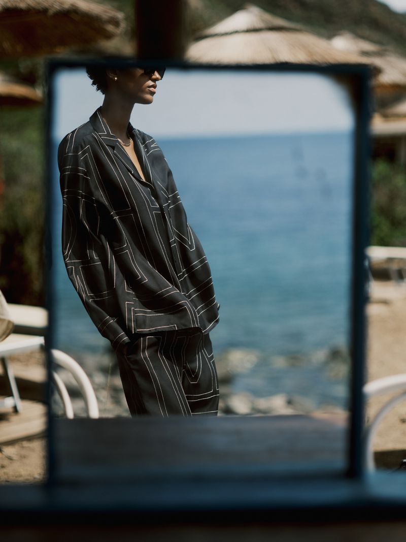 Woman in stylish patterned suit reflects in outdoor mirror against blue ocean backdrop, capturing a blend of fashion and serene scenery.