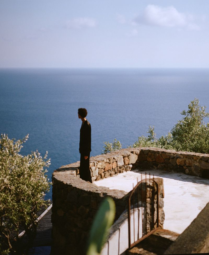 Adult stands on stone terrace overlooking the blue sea and sky, with green trees and other vegetation visible in the foreground.