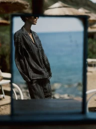 Reflection in a mirror of a model wearing a black patterned silk shirt and matching trousers with an ocean background.