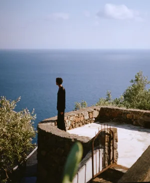Model standing on a stone terrace overlooking the blue ocean under a clear sky for Toteme Filicudi