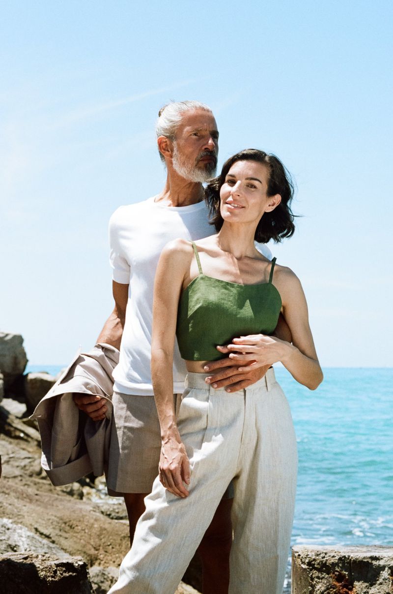 A man and woman in casual summer clothing pose near the ocean on a sunny day. The woman is wearing a green top and the man is in white.