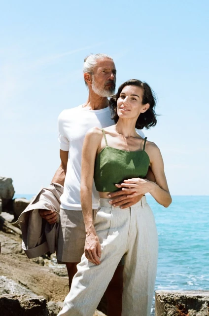 A man and woman in casual summer clothing pose near the ocean on a sunny day. The woman is wearing a green top and the man is in white.