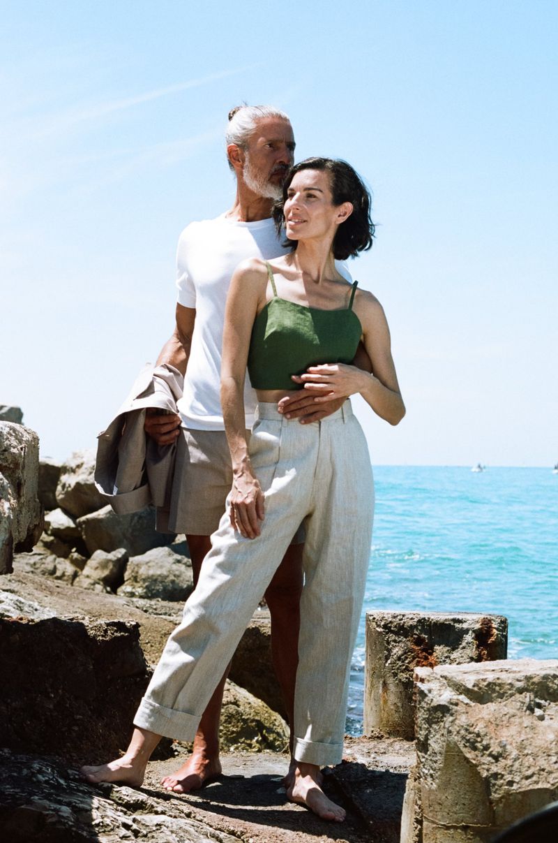 Mature couple stands on a stone wall overlooking the sea on a sunny day. The woman wears a green top and cream pants; the man wears white.