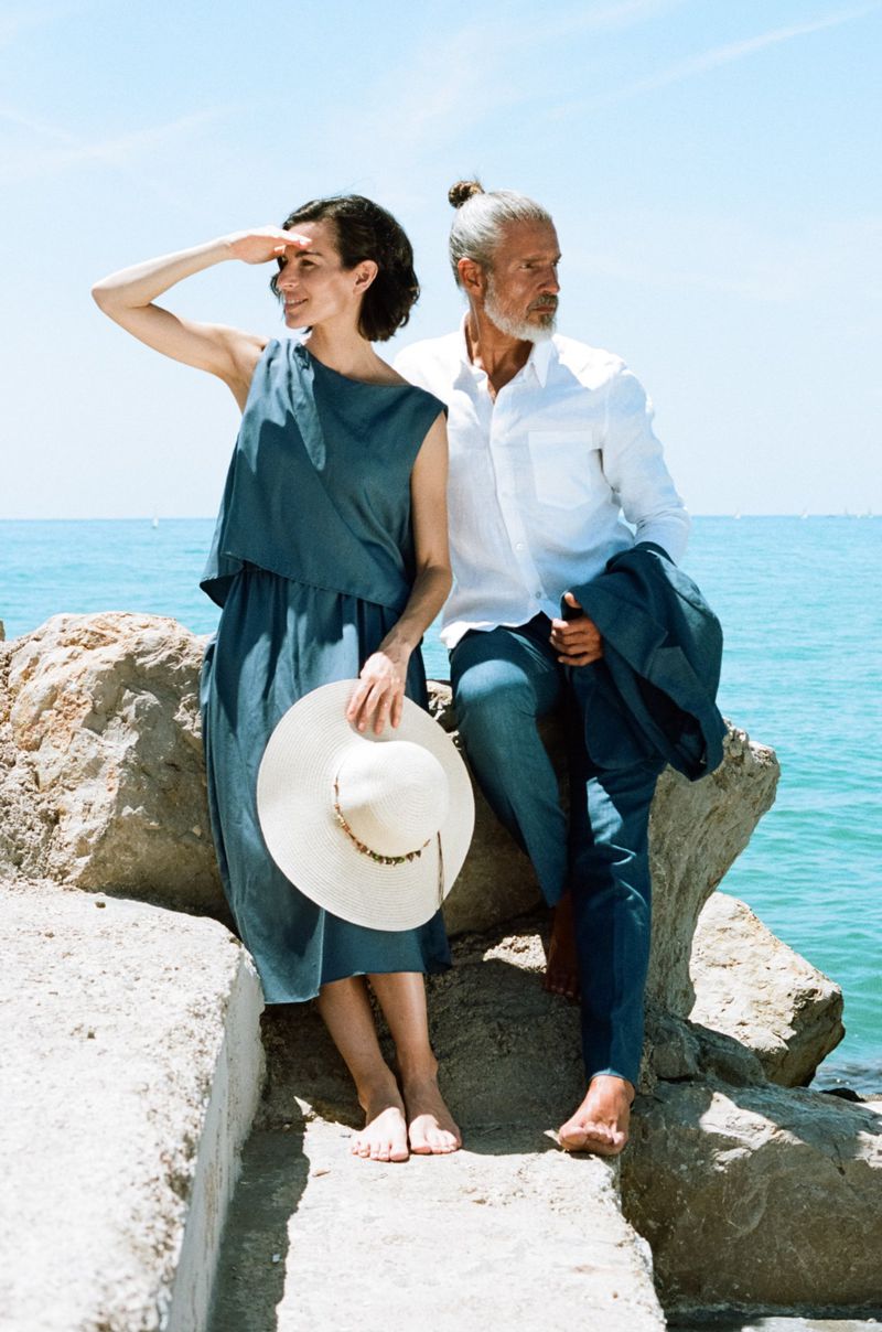 Mature couple posing on stone steps overlooking the turquoise ocean. Woman wears a blue dress and hat; man wears a white shirt and blue pants.
