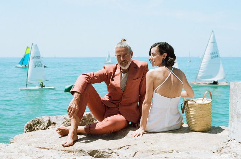 Mature couple poses on rocks by the sea, sun shining. The stylish pair are dressed in fashionable summer clothes with sailboats in the background.