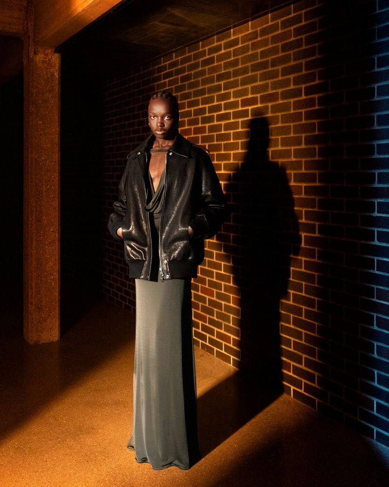 Fashion editorial shot of a Black woman in a leather jacket and long skirt standing against a textured brick wall, studio lighting.