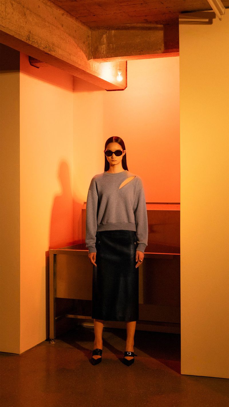 A woman wearing a grey sweater and black skirt stands indoors under orange lighting, posing in front of a shelf.