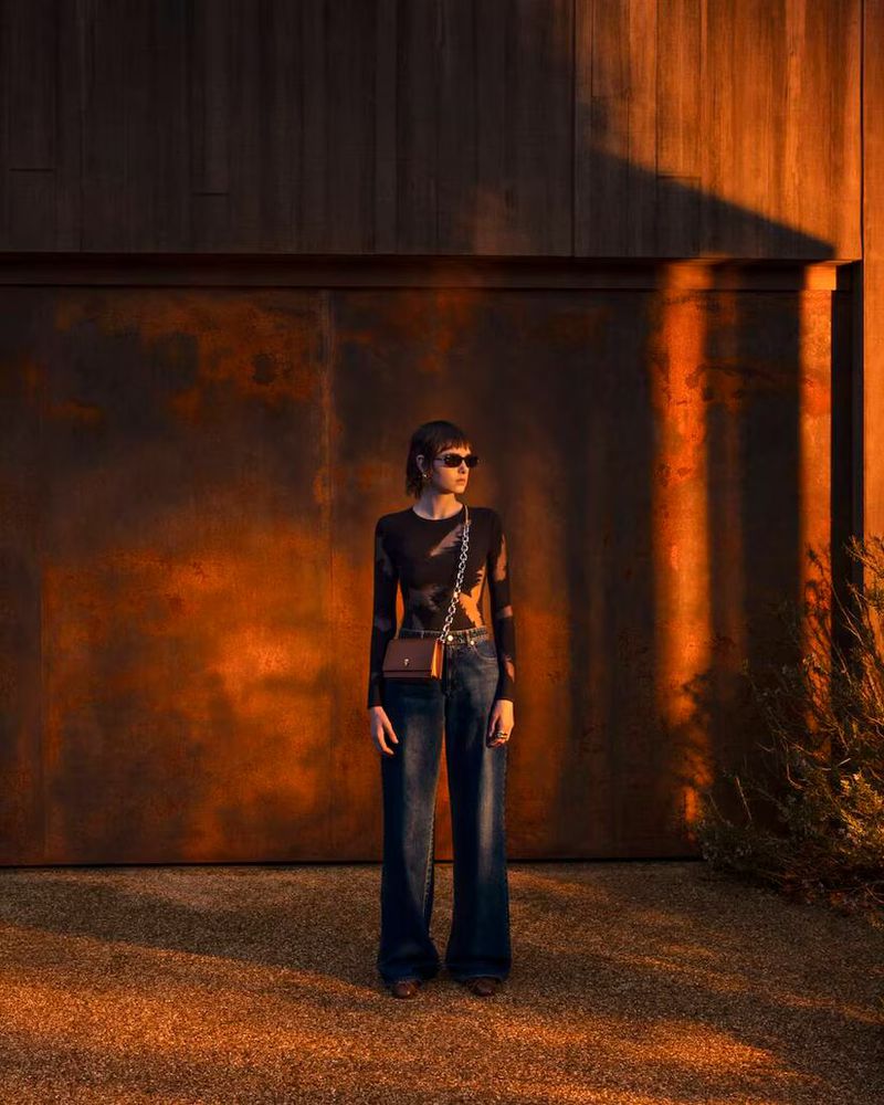 An adult female stands in front of a wooden wall in outdoor fashion shot. She wears dark jeans and a long-sleeved shirt with sunglasses.