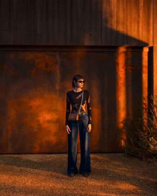 An adult female stands in front of a wooden wall in outdoor fashion shot. She wears dark jeans and a long-sleeved shirt with sunglasses.
