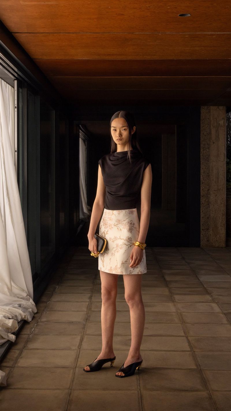 A full shot of a slender, light-skinned woman in a black top and patterned mini skirt, holding a clutch, indoors with a tiled floor.