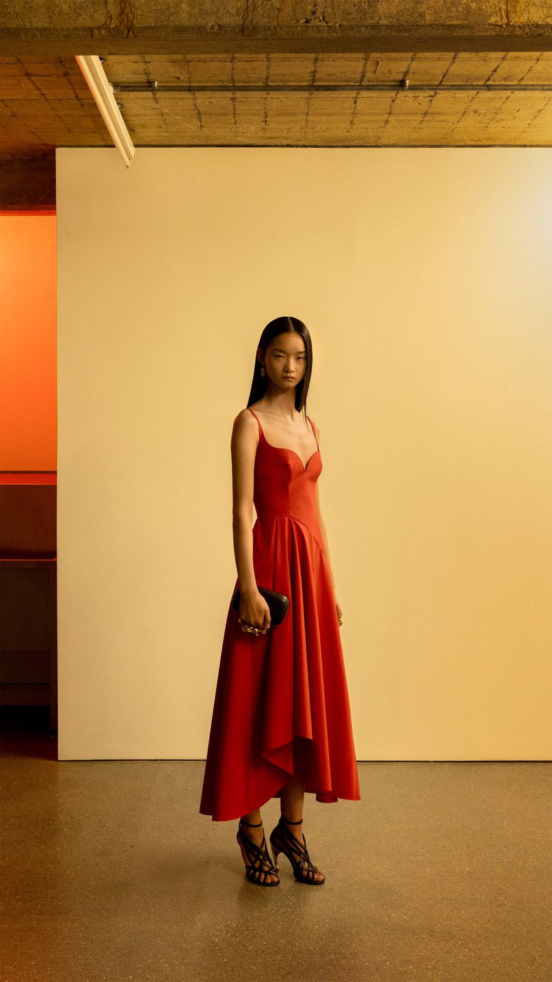 Elegant full-length portrait: A stylish Asian woman in a red dress holds a black clutch, standing in an urban setting.