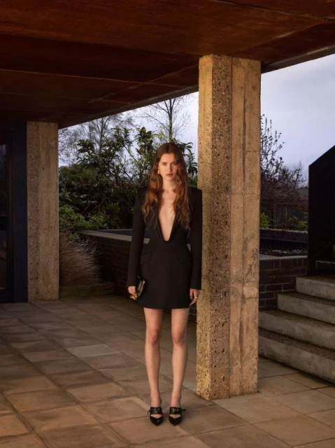 A woman in a black mini dress with a plunging neckline poses in a covered outdoor setting with concrete pillars and greenery in the background.