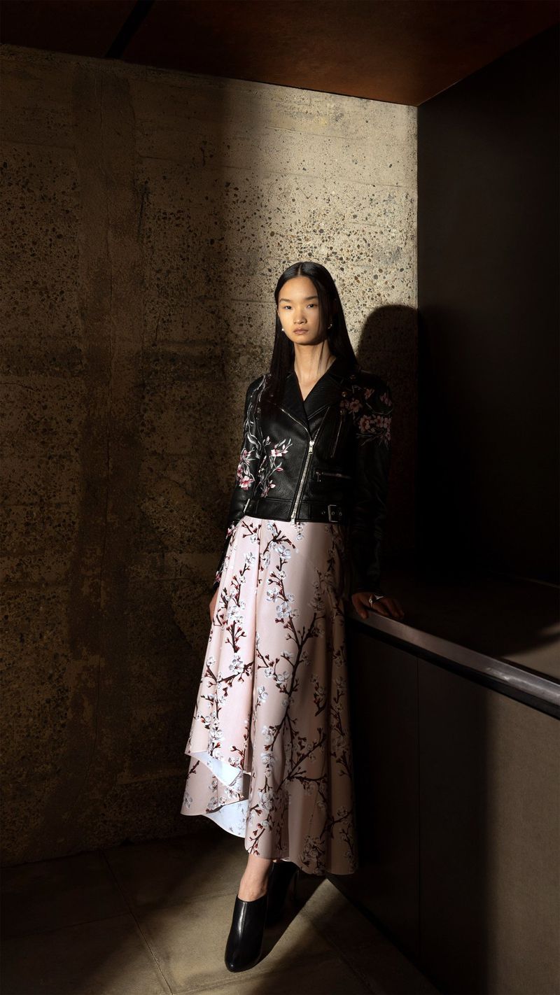 Asian woman in a black leather jacket and pale pink floral skirt poses in a dramatically lit interior setting with concrete walls.