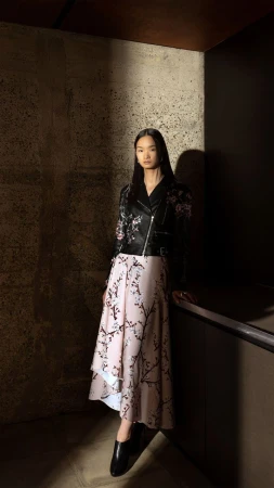 Asian woman in a black leather jacket and pale pink floral skirt poses in a dramatically lit interior setting with concrete walls.