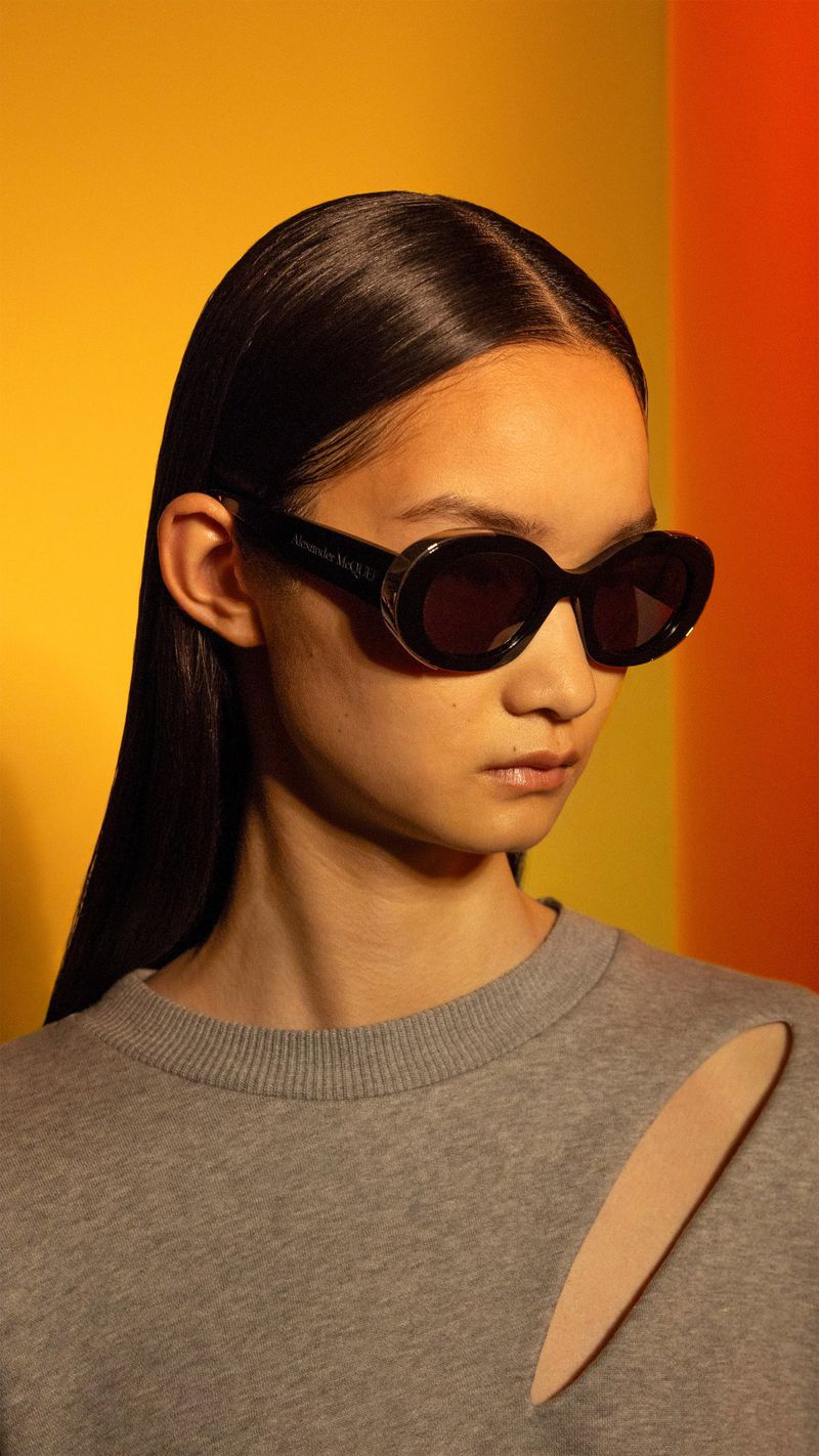 East Asian model wearing black oval sunglasses and a grey cutout sweatshirt against a warm yellow and orange backdrop.