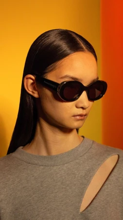 East Asian model wearing black oval sunglasses and a grey cutout sweatshirt against a warm yellow and orange backdrop.