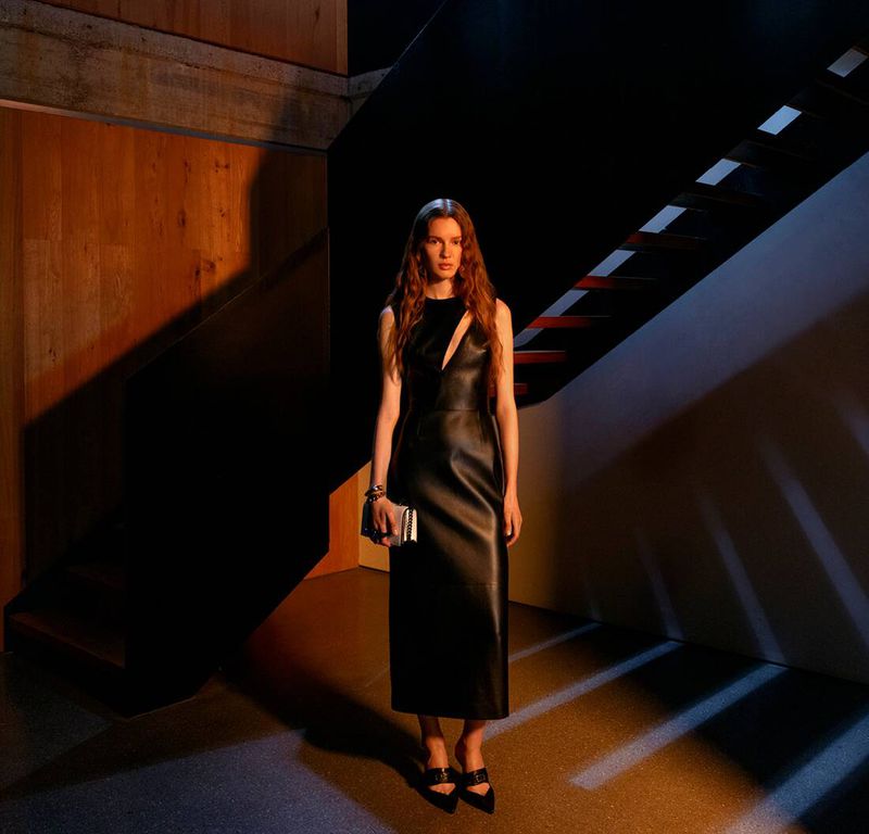 Model wearing a black leather cutout midi dress and black heels, holding a silver clutch beneath an architectural staircase.