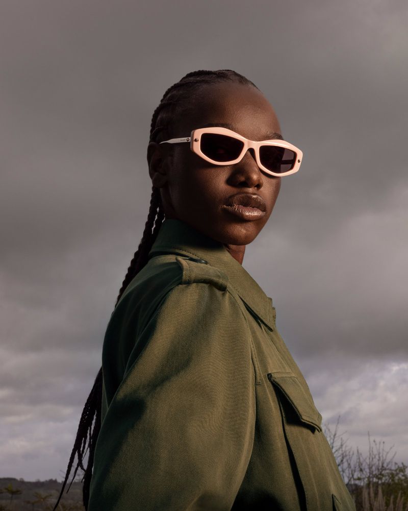 Woman wearing pink sunglasses and green jacket poses outdoors against a cloudy sky; braided hair is pulled back from her face.