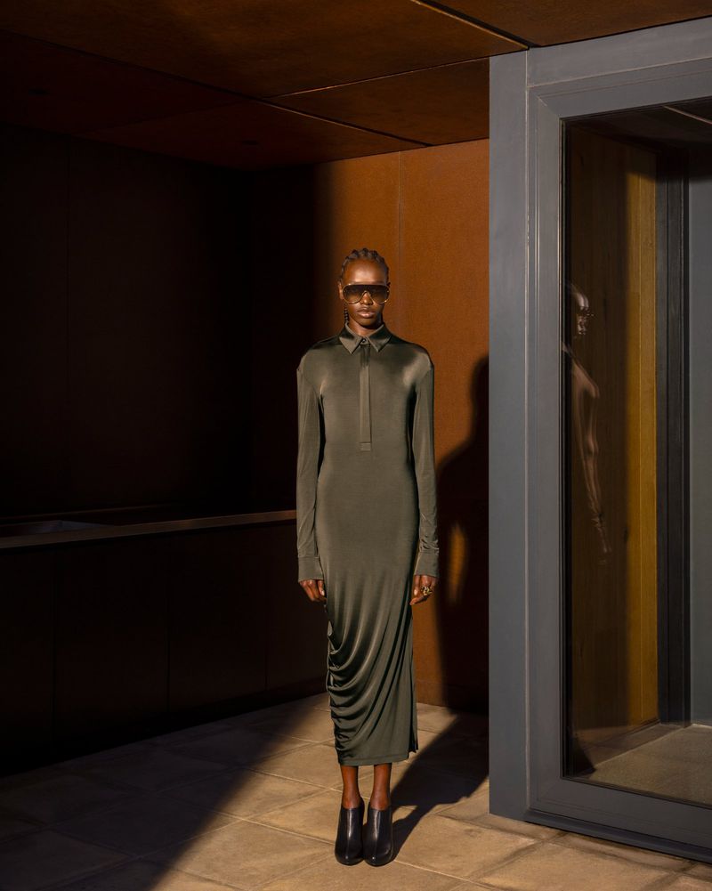 A slender woman poses in a long, olive-green dress and black boots. The space features brown walls and a concrete floor, softly illuminated.