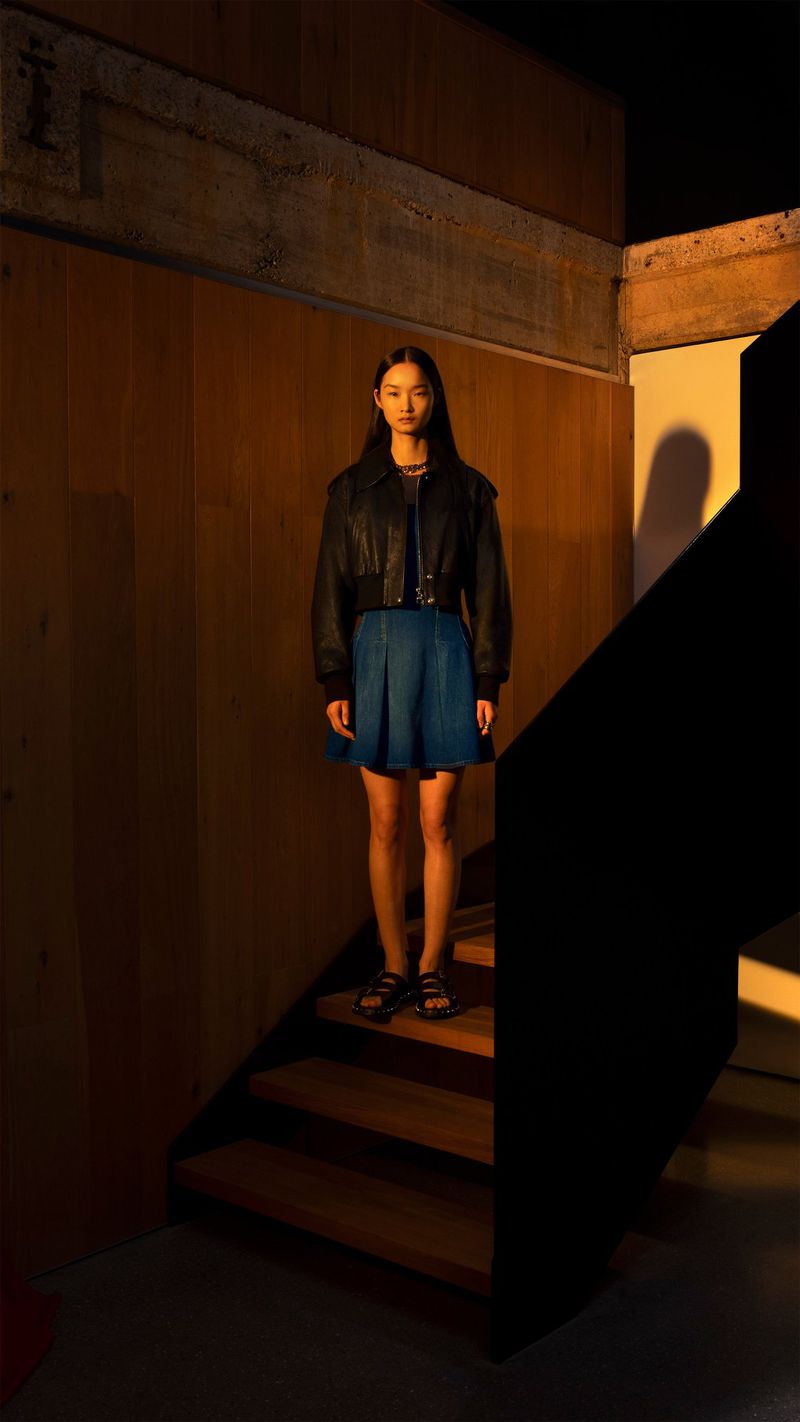 Young woman in a dark jacket and blue skirt stands on wooden stairs indoors in a moody fashion shot with warm lighting and shadows.