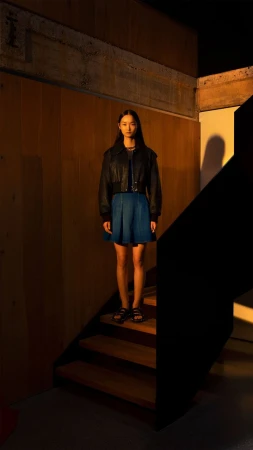 Young woman in a dark jacket and blue skirt stands on wooden stairs indoors in a moody fashion shot with warm lighting and shadows.