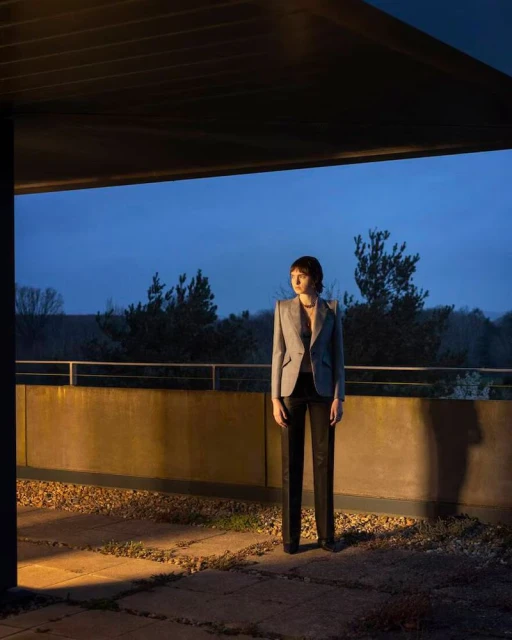 Adult female stands on a stone balcony outside at dusk. She wears a grey blazer, dark pants, and dark shoes near green trees.
