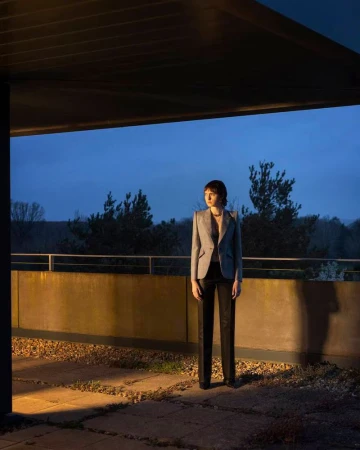 Adult female stands on a stone balcony outside at dusk. She wears a grey blazer, dark pants, and dark shoes near green trees.