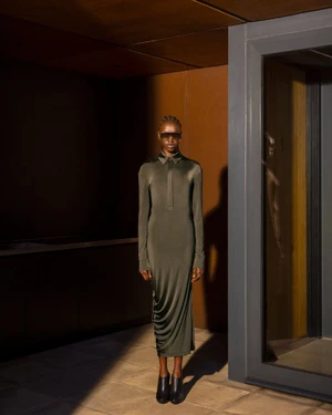 Model standing in a long dark green dress wearing sunglasses inside a building with warm lighting.
