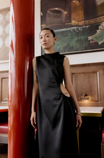 Elegant woman in a sleeveless black dress poses in an indoor setting with a red column and artwork in the background.