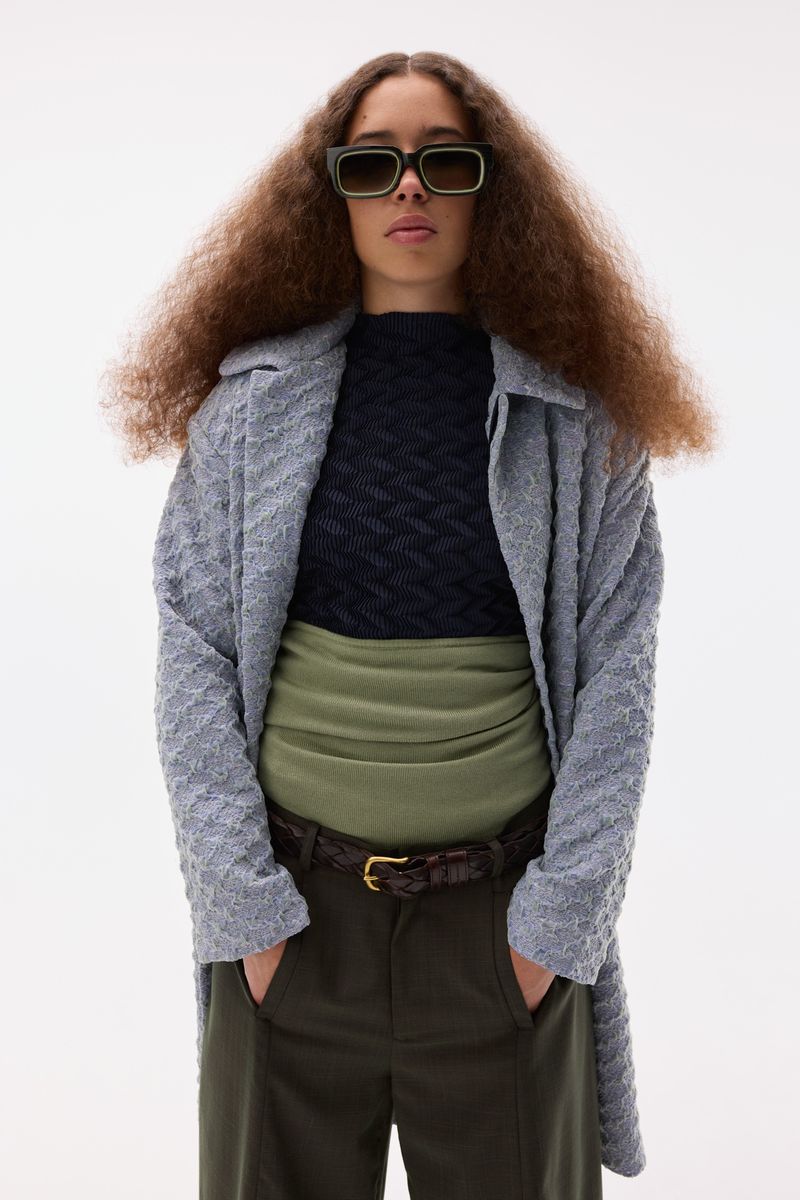 Young adult with curly hair poses in a photo studio wearing stylish sunglasses, sweater, trousers, coat, and belt in a fashion shot.