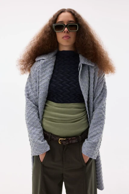 Young adult with curly hair poses in a photo studio wearing stylish sunglasses, sweater, trousers, coat, and belt in a fashion shot.
