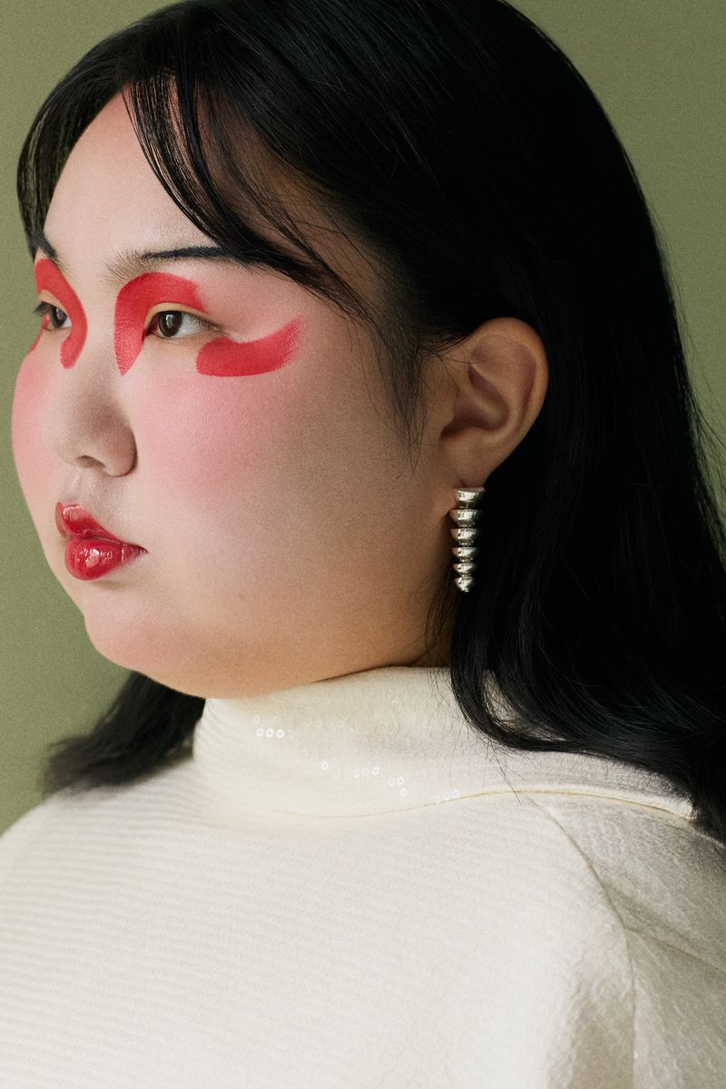 Close-up of an Asian woman with striking red makeup around her eyes, glossy red lips, and silver earrings, wearing an off-white top.