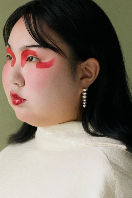 Close-up of an Asian woman with striking red makeup around her eyes, glossy red lips, and silver earrings, wearing an off-white top.