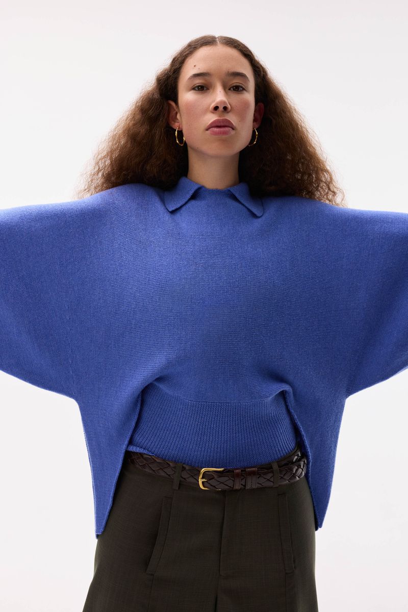 A woman in a blue sweater and olive pants with a woven belt is centered in a bright studio. Soft light emphasizes the texture of the sweater.