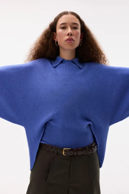 A woman in a blue sweater and olive pants with a woven belt is centered in a bright studio. Soft light emphasizes the texture of the sweater.