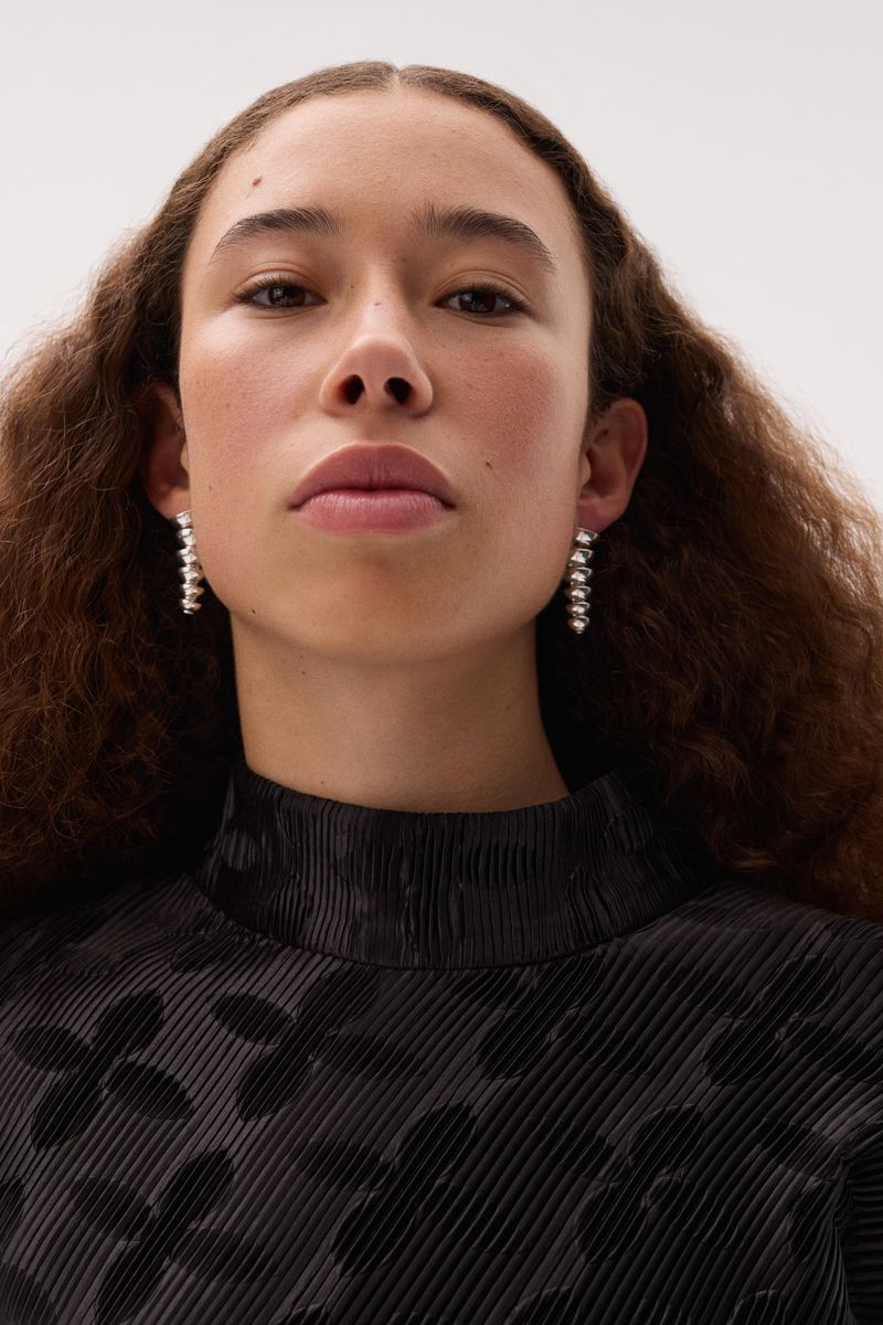 Portrait of a woman wearing silver earrings and a black floral patterned top, captured in a well-lit, close-up shot against a plain white background.
