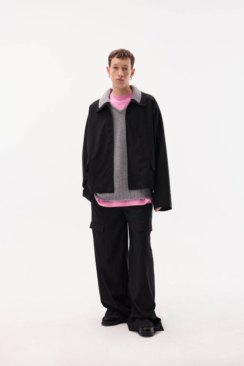 Full-length studio shot of a young adult male model wearing a black jacket, vest, pink top, and black pants against a white background.